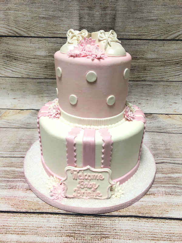 2 tier pink and white baby shower cake for a girl. with chocolate booties on top, white polka dots around top tier and pink and white strips on bottom layer. welcome baby plaque in front at bottom. 